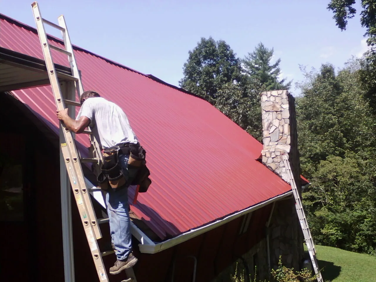 Roofer on a ladder beside a red metal roof and stone chimney — Soffit & Fascia Repair by Roofing Brothers in Starlight